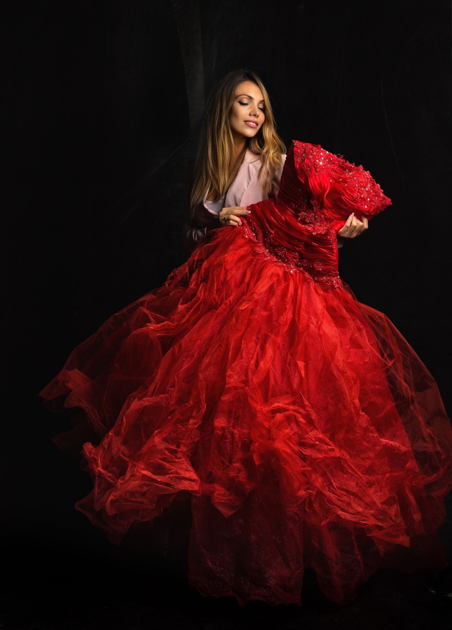 A woman wearing a voluminous red ball gown with a sheer tulle skirt and a pale pink top, posed against a dark black background.